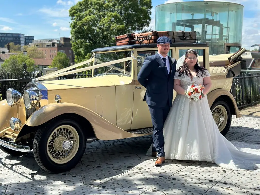 Bride and groom posing beside vintage gold wedding car with ribbon and suitcases on roof, wedding cars Norwich by Celeb Events.